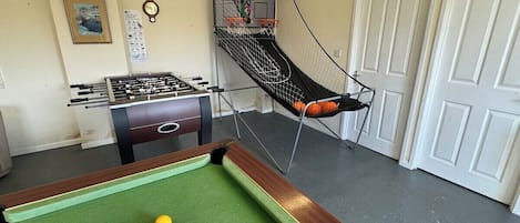 Mesa de bilhar com bolas, mesa de futebol de mesa e um aro de basquete em um ambiente com portas brancas.