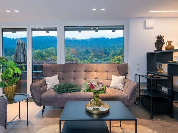 Un salon moderne avec un canapé, une table basse et une vue sur un paysage boisé, visible à travers de grandes fenêtres.
