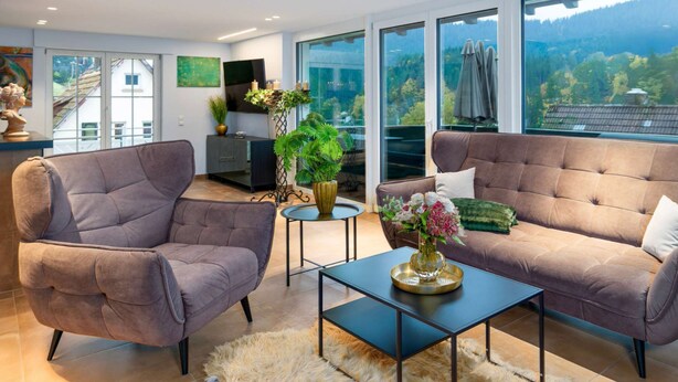 Una sala moderna con dos sofás, una mesa de centro y un televisor sobre un mueble bajo. Hay ventanas amplias con vistas a una zona boscosa.