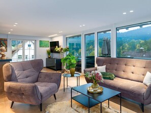 A modern living room with two sofas, a coffee table, and a TV on a stand. There are large windows with a view of a forested area. - Waldklang - Panorama | table football | smarthome | sauna | fenced | 10 persons (Alpirsbach)