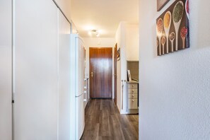Un pasillo estrecho con una puerta de madera al final, un refrigerador blanco a la izquierda y una zona de cocina con encimera y armarios a la derecha.