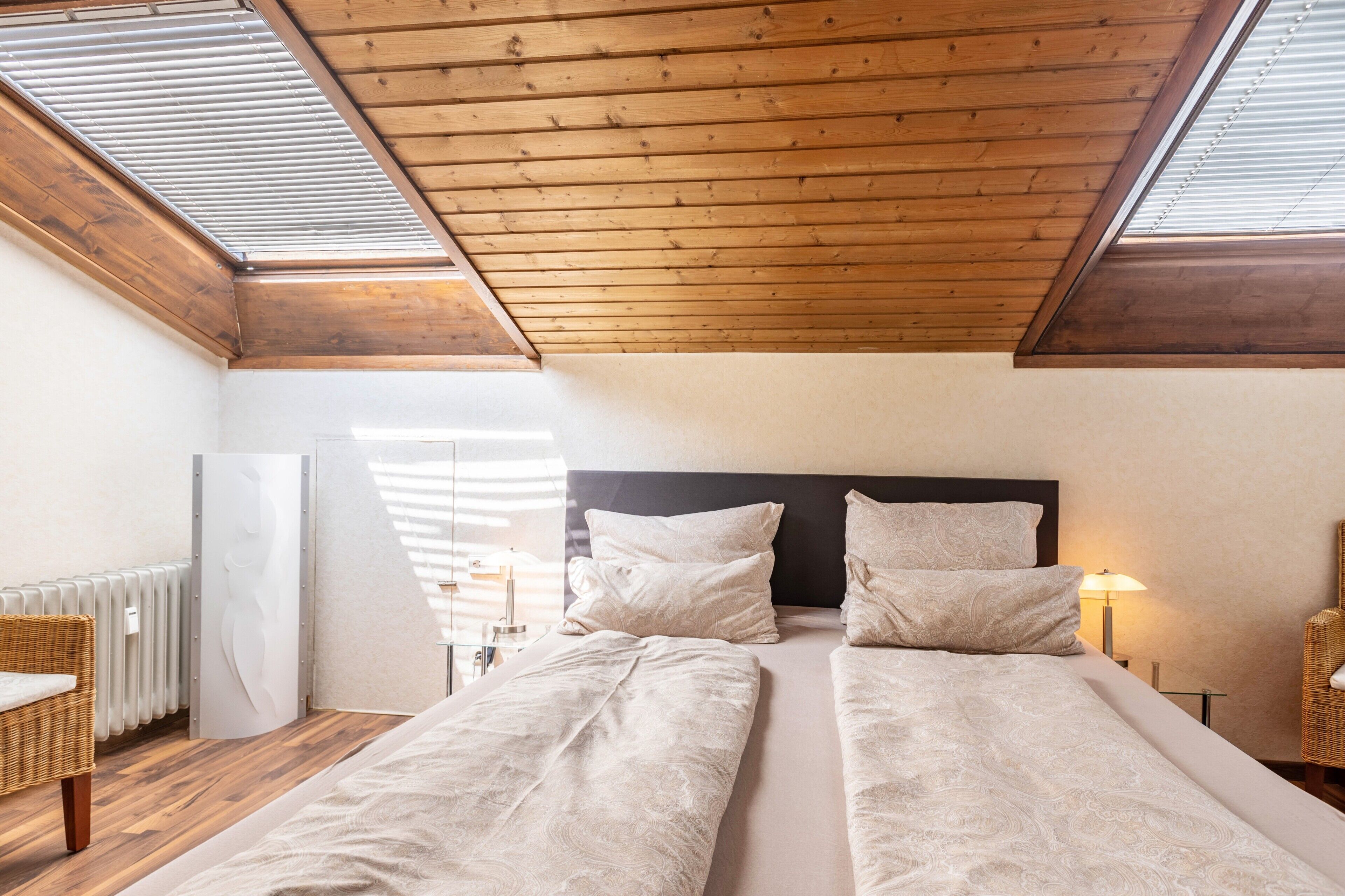Une chambre avec deux lits, des poutres apparentes en bois au plafond, des fenêtres de toit, un radiateur et des meubles en osier.