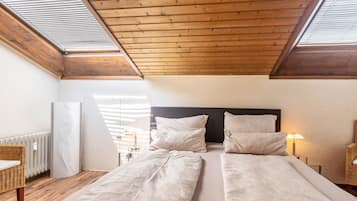 Une chambre avec deux lits, des poutres apparentes en bois au plafond, des fenêtres de toit, un radiateur et des meubles en osier.