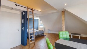 Une chambre aménagée en mezzanine avec un lit, une chaise verte, une table, une échelle et une fenêtre.