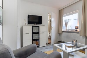 A modern living room with a flat-screen TV, a glass coffee table, a sofa, and a walk-in closet. - Bungalow 'Leona' with Private Terrace, Shared Garden and Wi-Fi (Büsum)