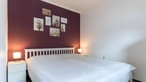 Uma cama bem arrumada com cabeceira branca, duas luminárias de cabeceira e uma parede com quadros.
