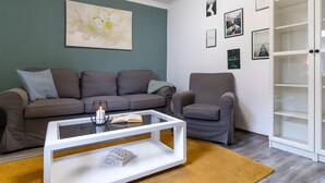 Uma sala de estar com sofá, poltrona, mesa de centro de vidro e estante de livros.