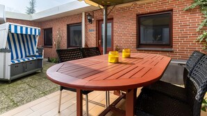 Um pátio externo com mesa redonda de madeira, duas cadeiras, um guarda-sol amarelo e um sofá azul listrado.