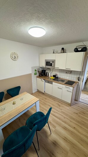 A compact kitchen with a microwave, sink, and cabinets. There is a dining table with chairs and a clock on the wall. - Bungalow 'Leona' with Private Terrace, Shared Garden and Wi-Fi (Büsum)