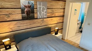 A bedroom with a wooden headboard, a bed with a patterned blue bedspread, two bedside tables with lamps, a rug, and a wall with a 'HOME' sign and a picture of a cow. - Apartment 'Malia', Shared Terrace and Wi-Fi (Büsum)