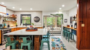 Een moderne keuken met een centraal eiland, groene barkrukken en een vloerkleed met patroon dat leidt naar een zithoek met een bank en stoelen.