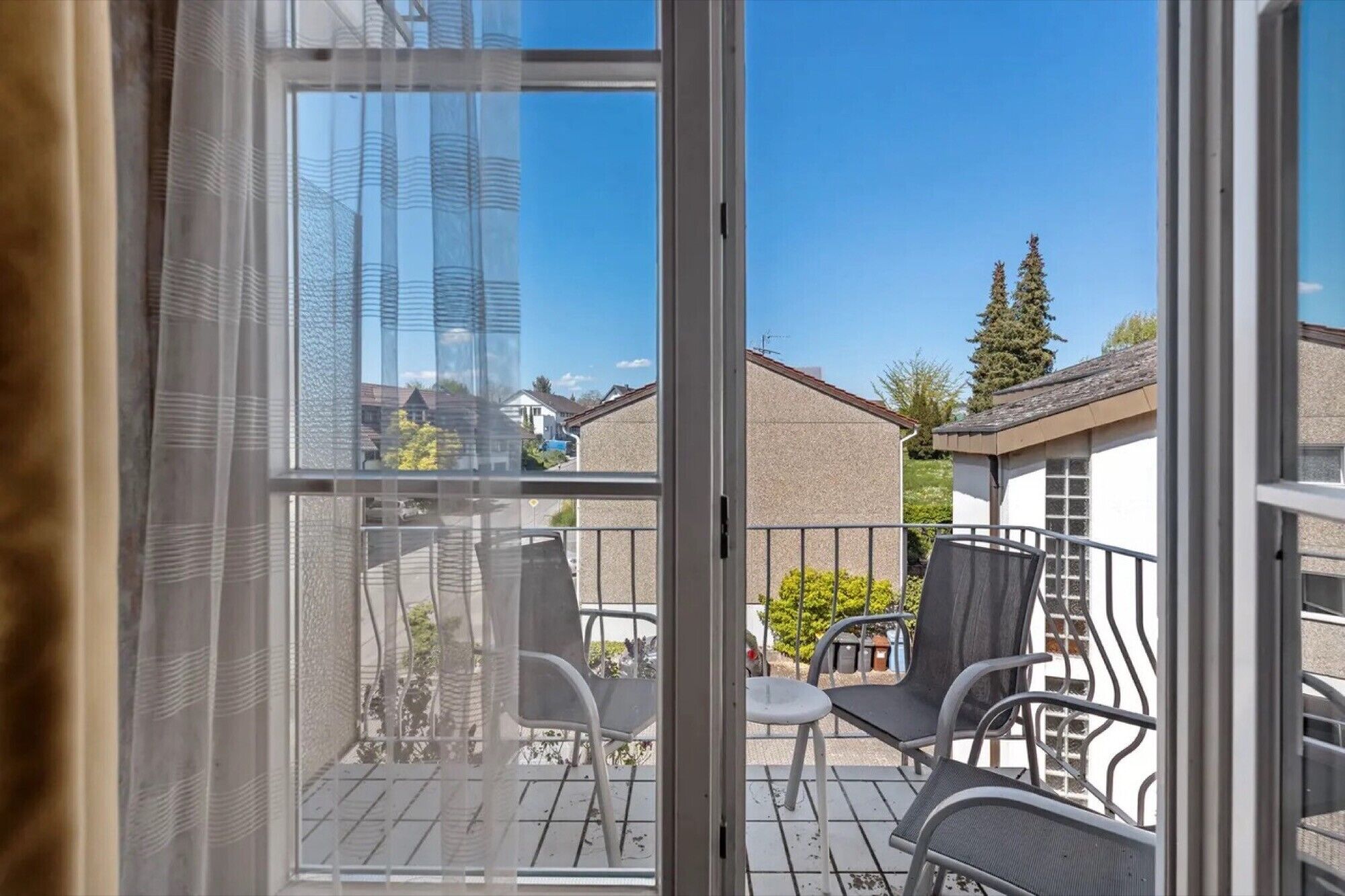 A balcony with chairs and a table, overlooking a residential area with houses and a tall building.