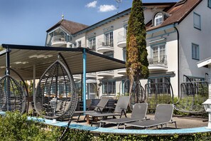 Un espace extérieur aménagé avec des meubles de jardin en osier, comprenant des chaises et des tables, un grand arbre et, en arrière-plan, un bâtiment doté de balcons.