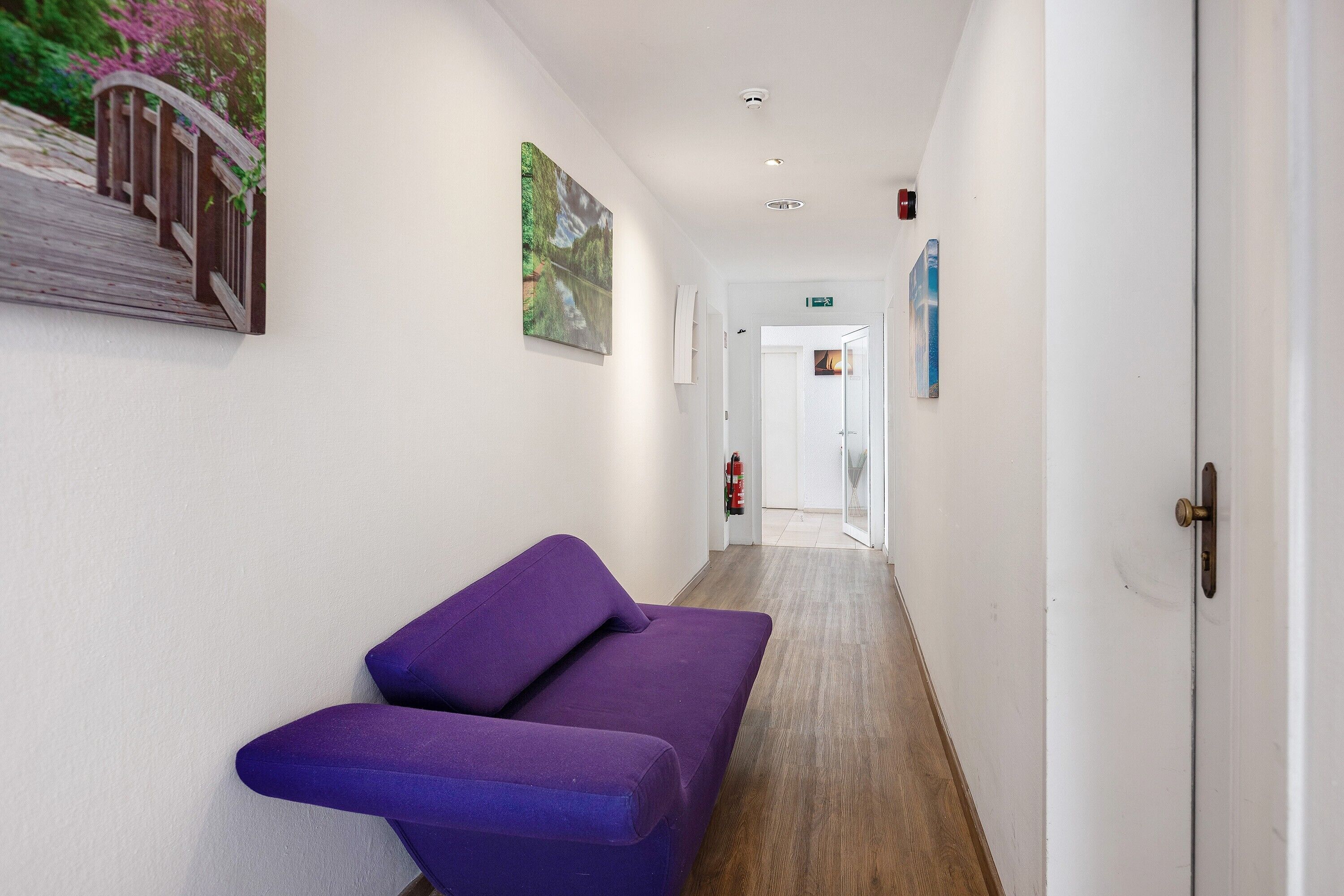 Un couloir avec un canapé violet, une œuvre d’art représentant un pont en bois et une porte donnant sur une pièce.