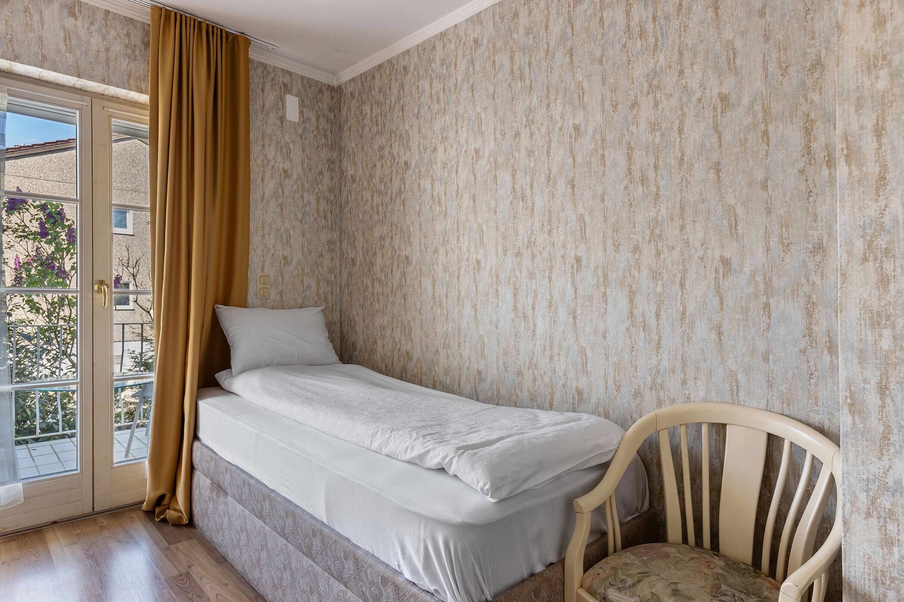 Uma cama de solteiro com cabeceira, uma cadeira, uma janela com cortinas e uma parede texturizada.