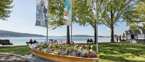 Un parque frente al agua con un macetero con forma de bote de madera lleno de flores, bancos y banderas.