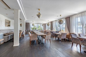Una àrea de dinar espaiosa amb taules llargues i cadires, grans finestres amb cortines i un sostre alt amb il·luminació decorativa.