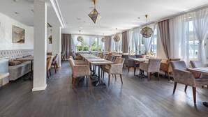 Un'ampia sala da pranzo con lunghe tavolate e sedie, ampie finestre con tende e un soffitto alto con illuminazione decorativa.