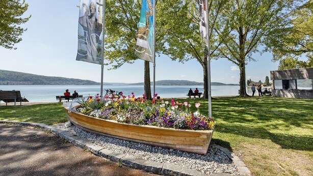 Un parco sul lungomare con un fioriere a forma di barca di legno, panchine e bandiere.