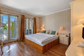Habitación de hotel con una cama grande, suelo de madera, una mesita de noche pequeña con lámpara y vistas a través de un ventanal.