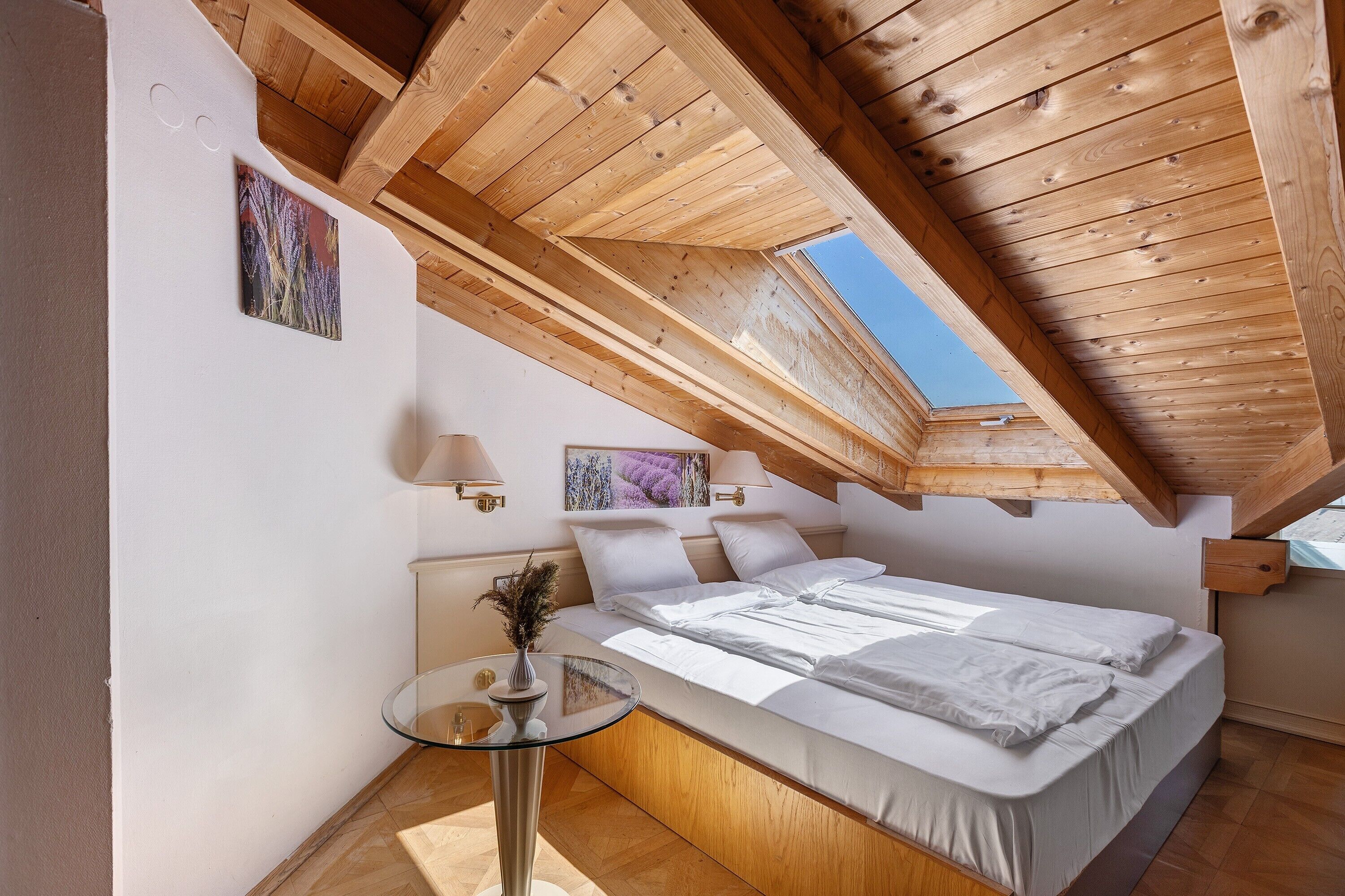 A bedroom with a sloped ceiling, a bed, a small table with a vase, and wall-mounted lamps.