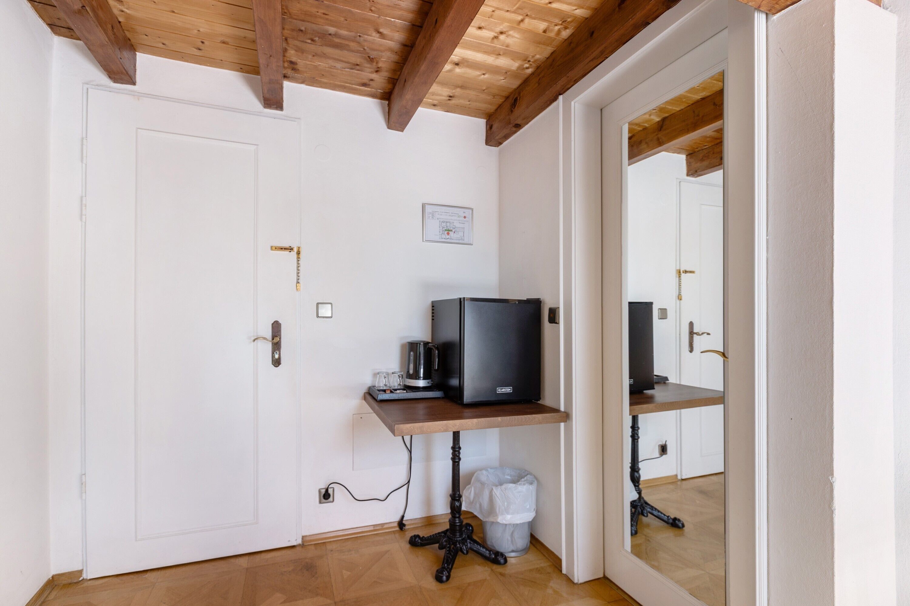 A room with a wooden ceiling, a white door, a small table with a black television, a trash bin, and a mirror reflecting the room.