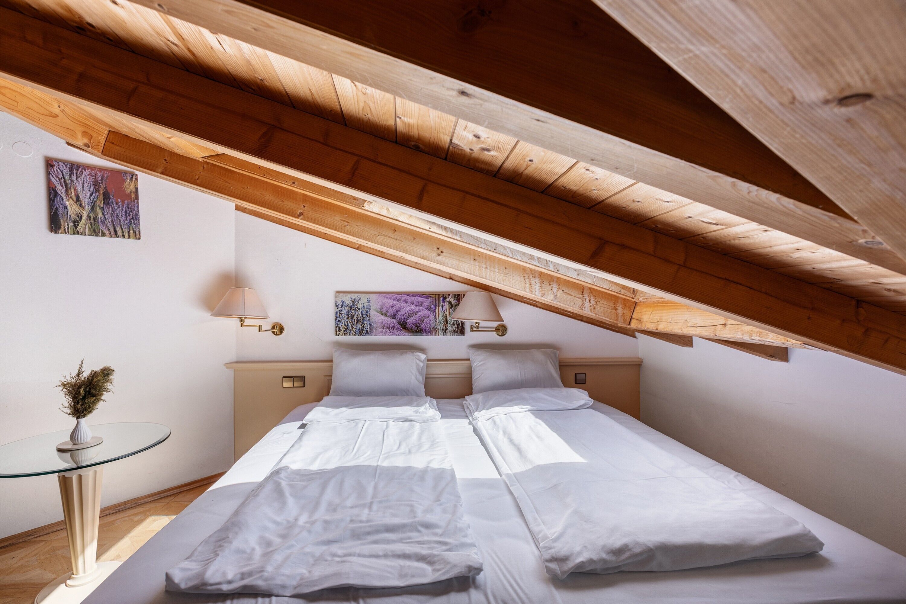 A bedroom with a sloped ceiling, a bed with white bedding, a bedside table with a lamp, and a wall-mounted artwork.