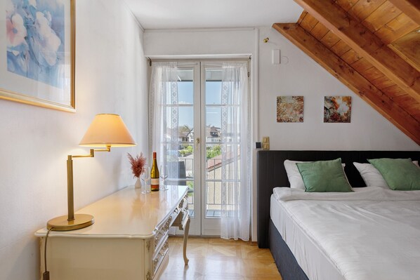 A bedroom with a bed, a desk with a lamp, a window with curtains, and artwork on the wall.
