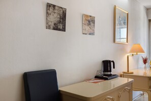 Ein Hotelzimmer mit Schreibtisch, Stuhl, Kaffeemaschine, Lampe und zwei gerahmten Kunstwerken an der Wand.