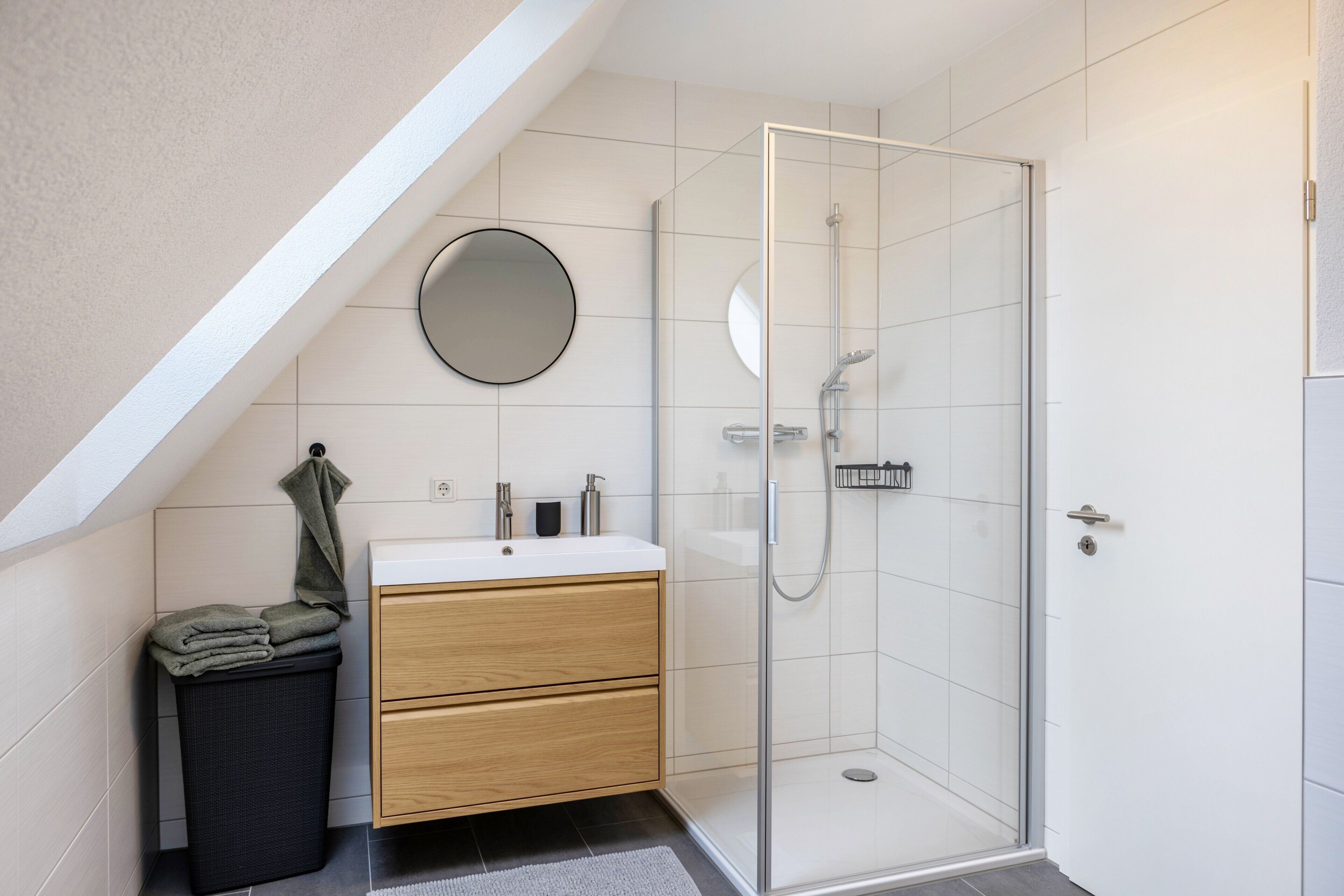 A modern bathroom with a glass shower enclosure, a wooden vanity, a round mirror, and a trash bin.