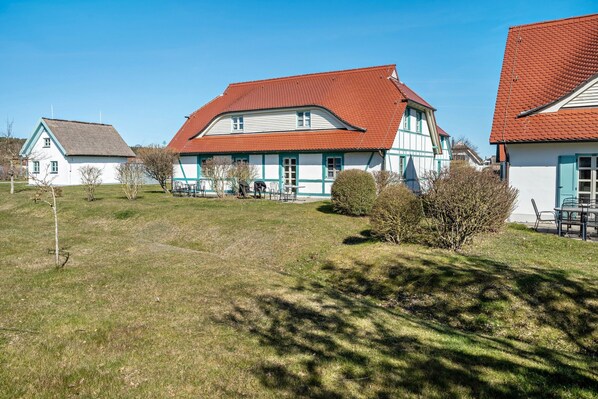 Une maison traditionnelle avec un toit rouge, des murs blancs et des bordures bleues, entourée d’une pelouse et d’arbres.