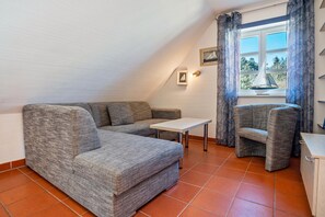 A small, cozy room with a grey sofa, a small table, and a chair. There is a window with curtains and a sailboat decoration. - Apartment 'B 53' with Wi-Fi (Dranske)
