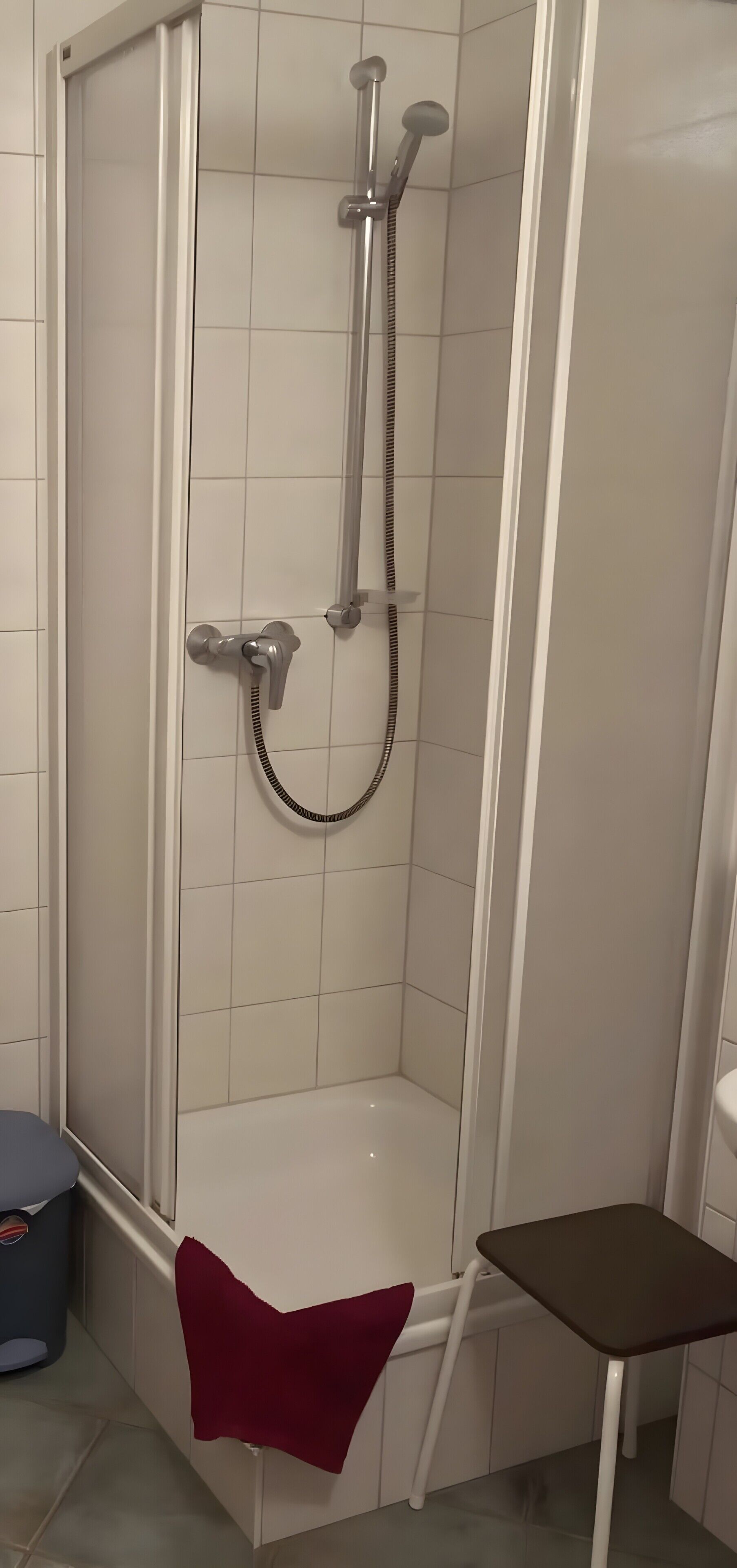 Eine Duschkabine mit Glastür, einer Handbrause und einem roten Handtuch auf dem Boden.