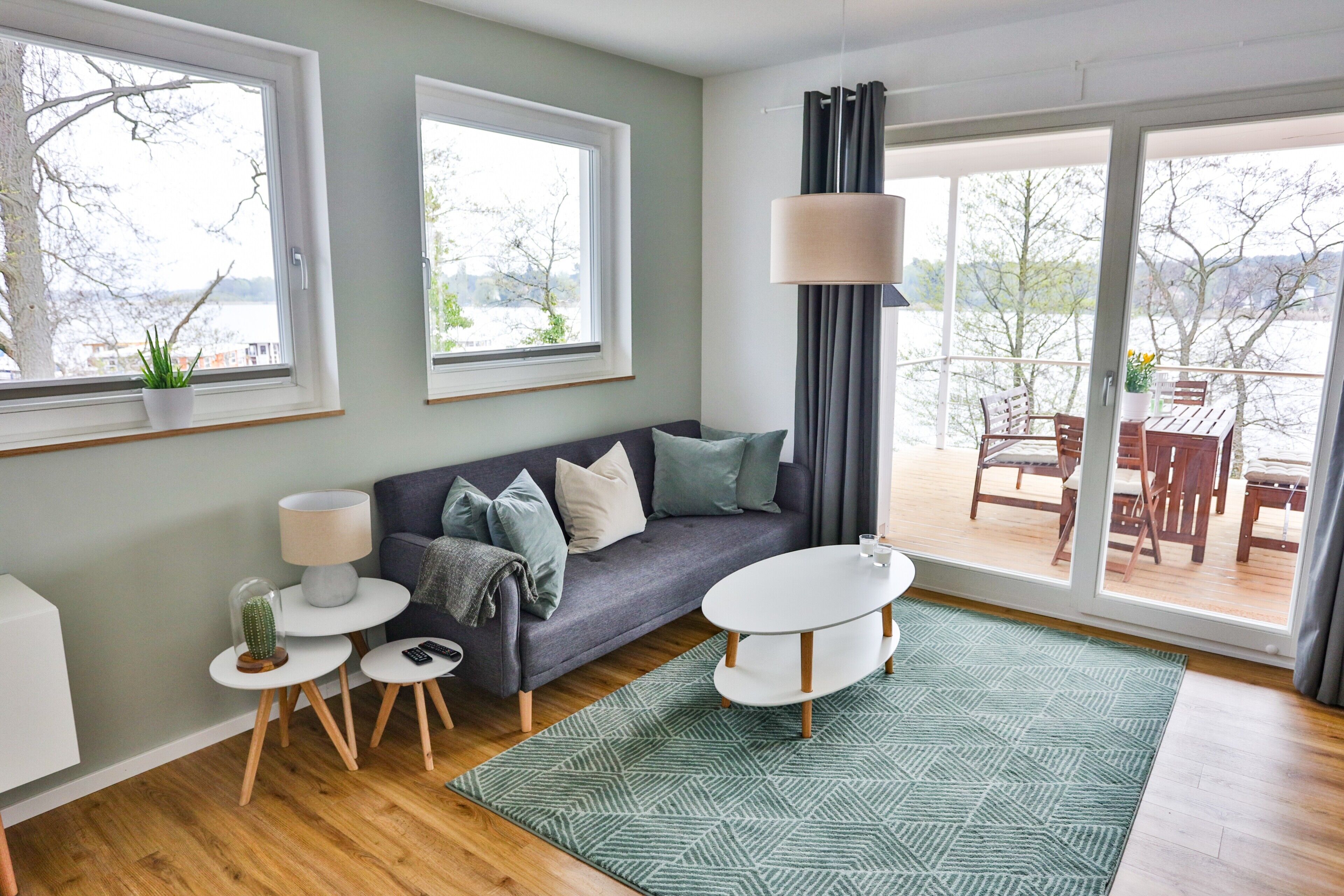 Ein modernes Wohnzimmer mit einer Couch, einem Couchtisch, einer Lampe, einem Kakteen und Blick ins Freie durch große Fenster.
