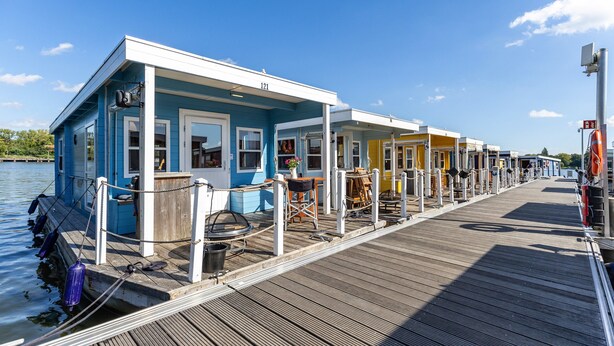 Une rangée de péniches colorées, dotées de terrasses et de garde-corps en bois, reliées par une jetée.