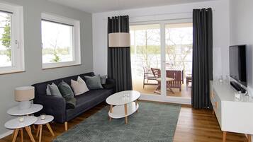A modern living room with a sofa, coffee table, and TV stand, large windows, and a sliding glass door to an outdoor area.