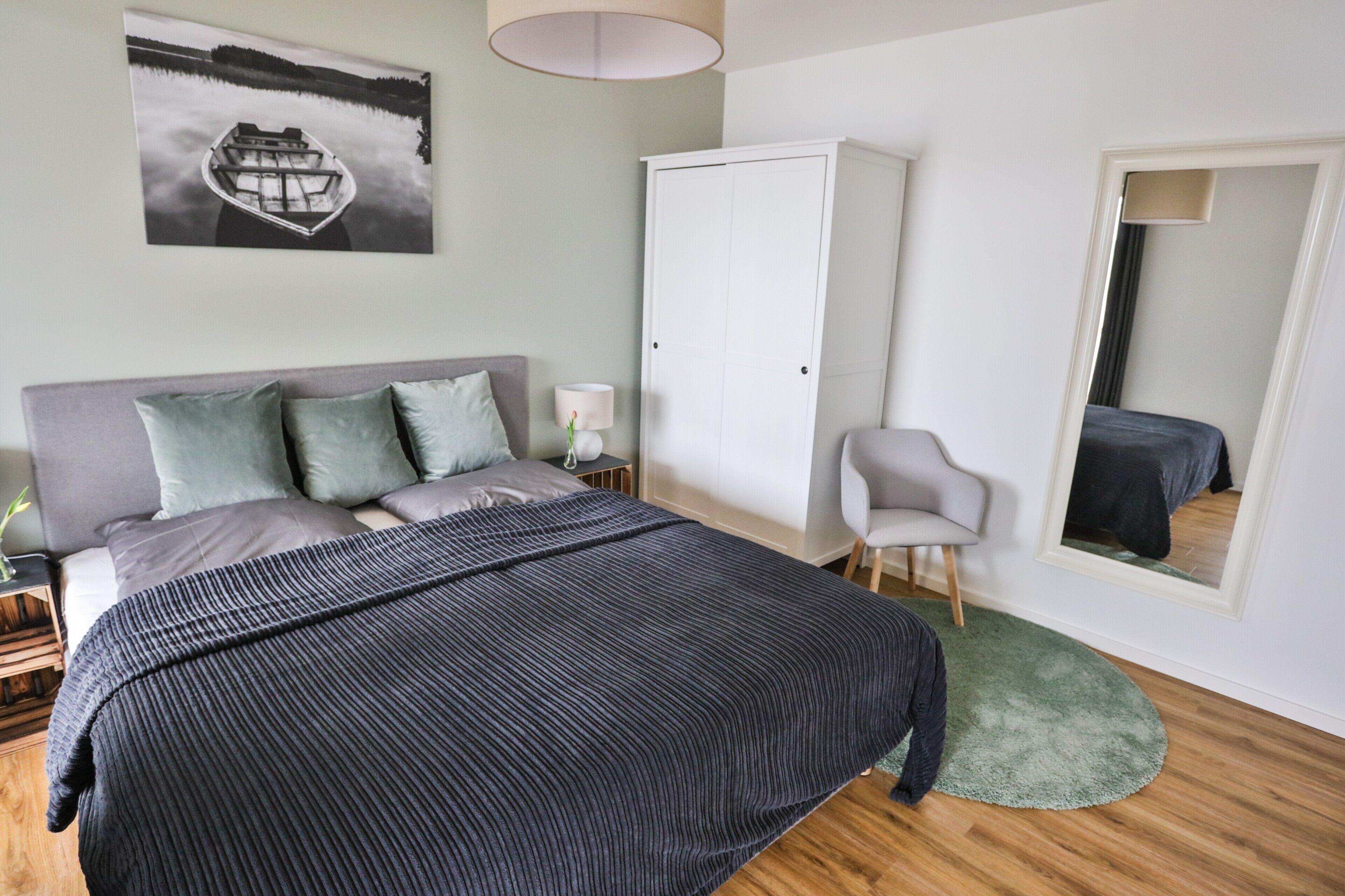 A bedroom with a bed, a wardrobe, a chair, a bedside table, a lamp, and a painting on the wall.