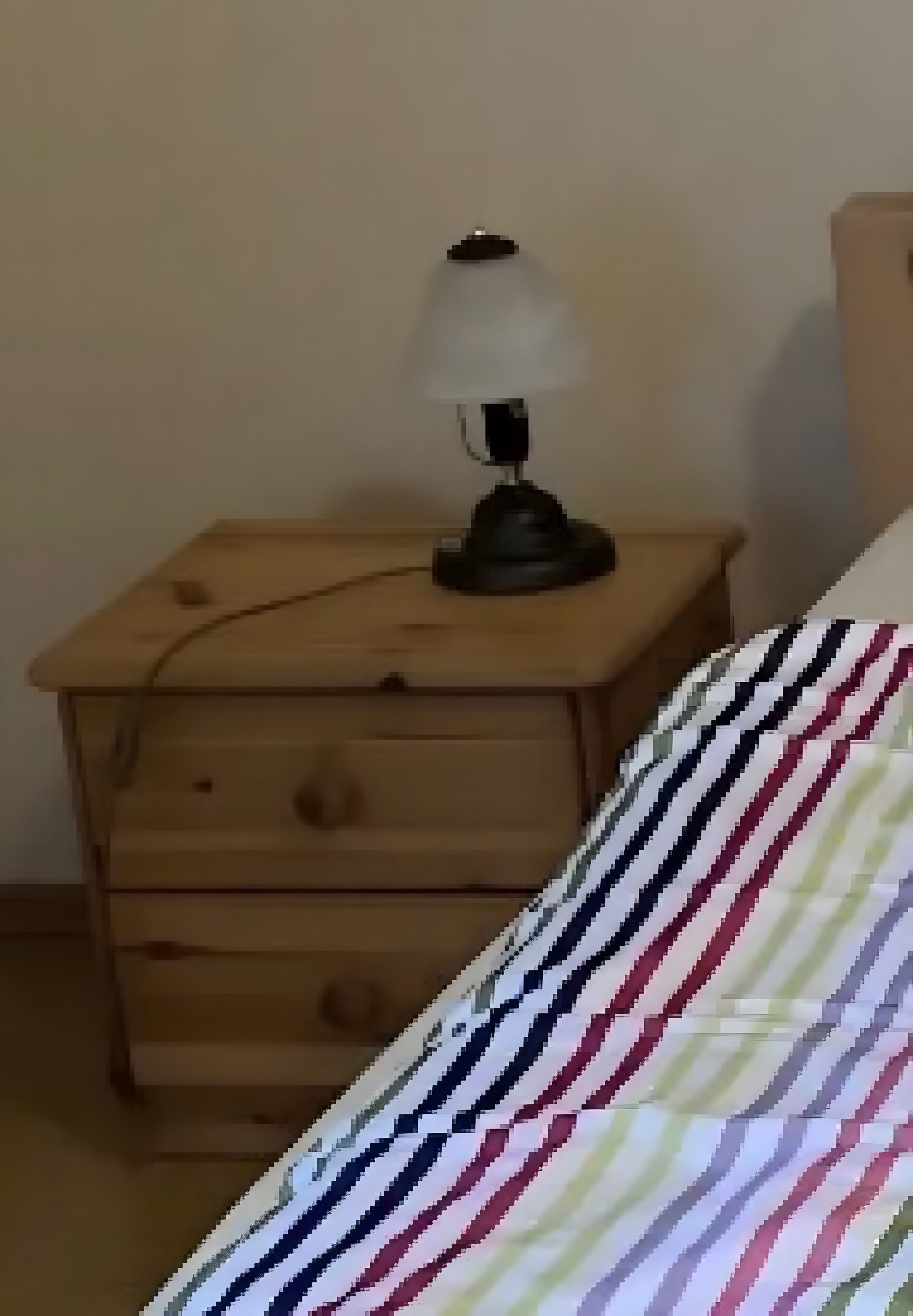 Une table de chevet en bois avec une lampe, un lit avec une literie à rayures et un mur uni.