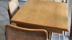Une petite table carrée en bois, avec deux chaises, une grille d’aération et une porte à l’arrière-plan.