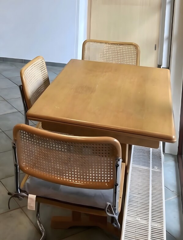 Une petite table carrée en bois, avec deux chaises, une grille d’aération et une porte à l’arrière-plan.