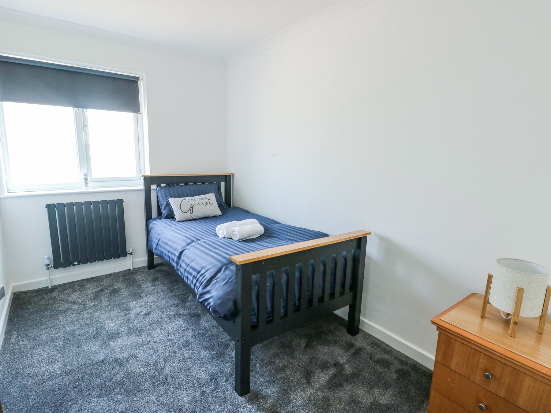 Une petite chambre bien rangée, avec un lit simple, un radiateur, une fenêtre et une commode en bois.