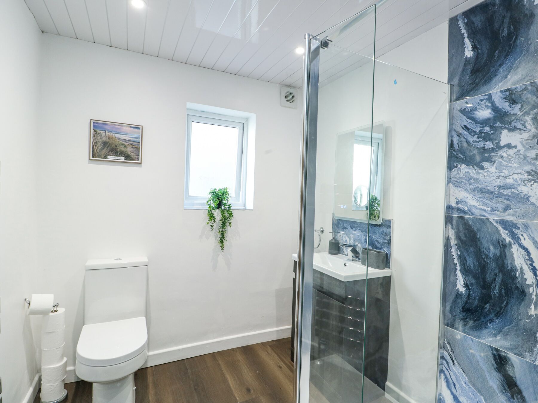 A modern bathroom with a toilet, a glass-enclosed shower, a sink, and a window with a plant.