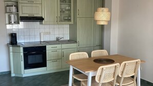 Una cocina con armarios verde claro, un horno negro y una zona de comedor con mesa de madera y sillas de mimbre.