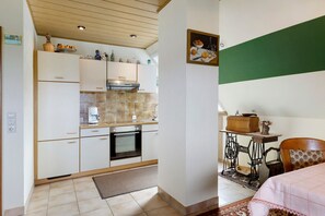 Cocina pequeña con estufa, horno y armarios blancos. Hay una mesita con una silla y una cama con colcha rosa.
