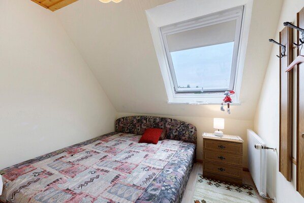 Un dormitorio con techo inclinado, una ventana, una cama con colcha estampada, una mesita de noche con lámpara y un radiador.