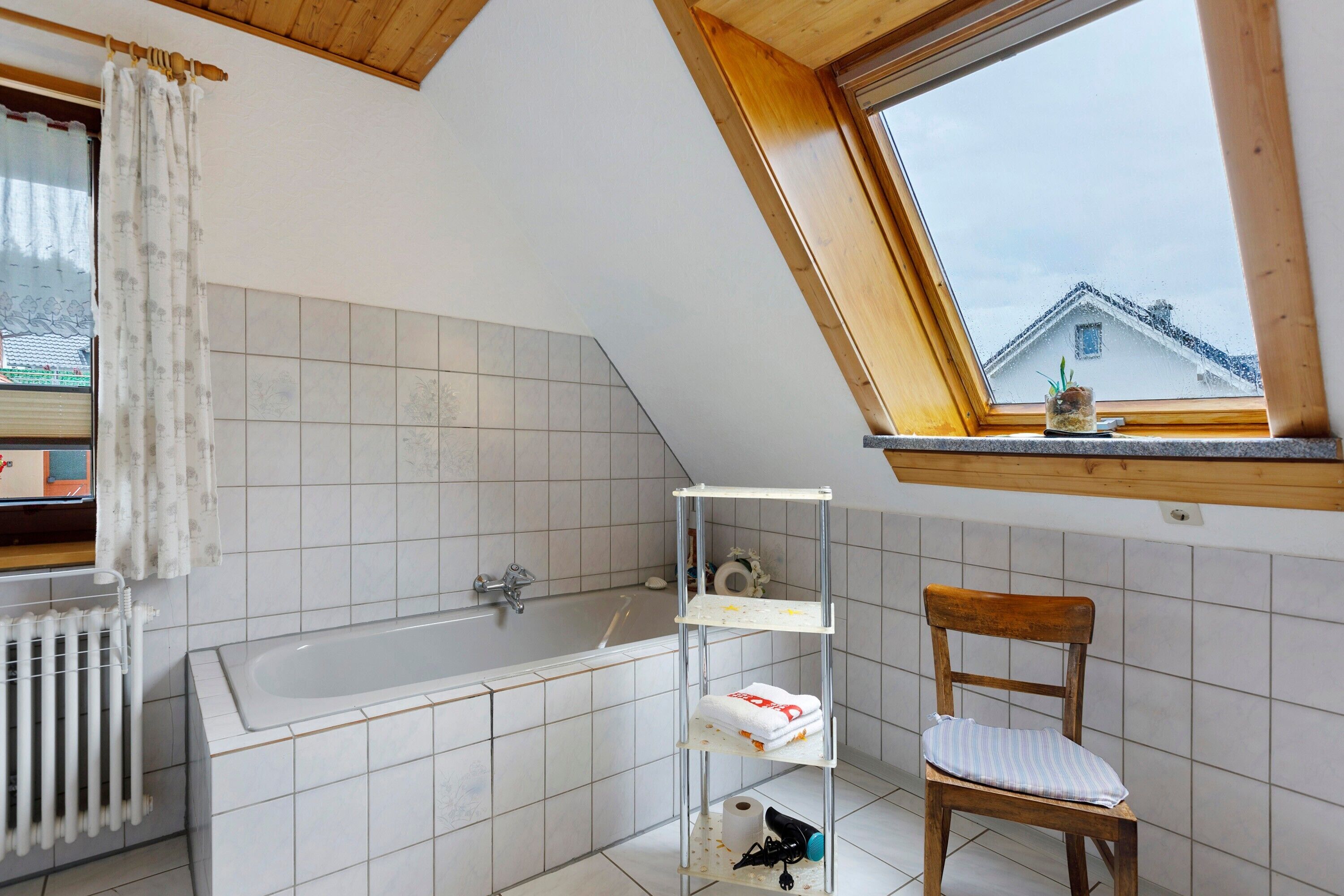 Een badkamer met een badkuip, een houten stoel, een plank met handdoeken en een raam met uitzicht op een huis.