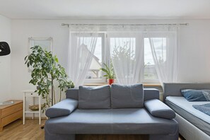 A modern living room with a grey sofa, a small white side table, a large potted plant, and a view through sheer curtains.