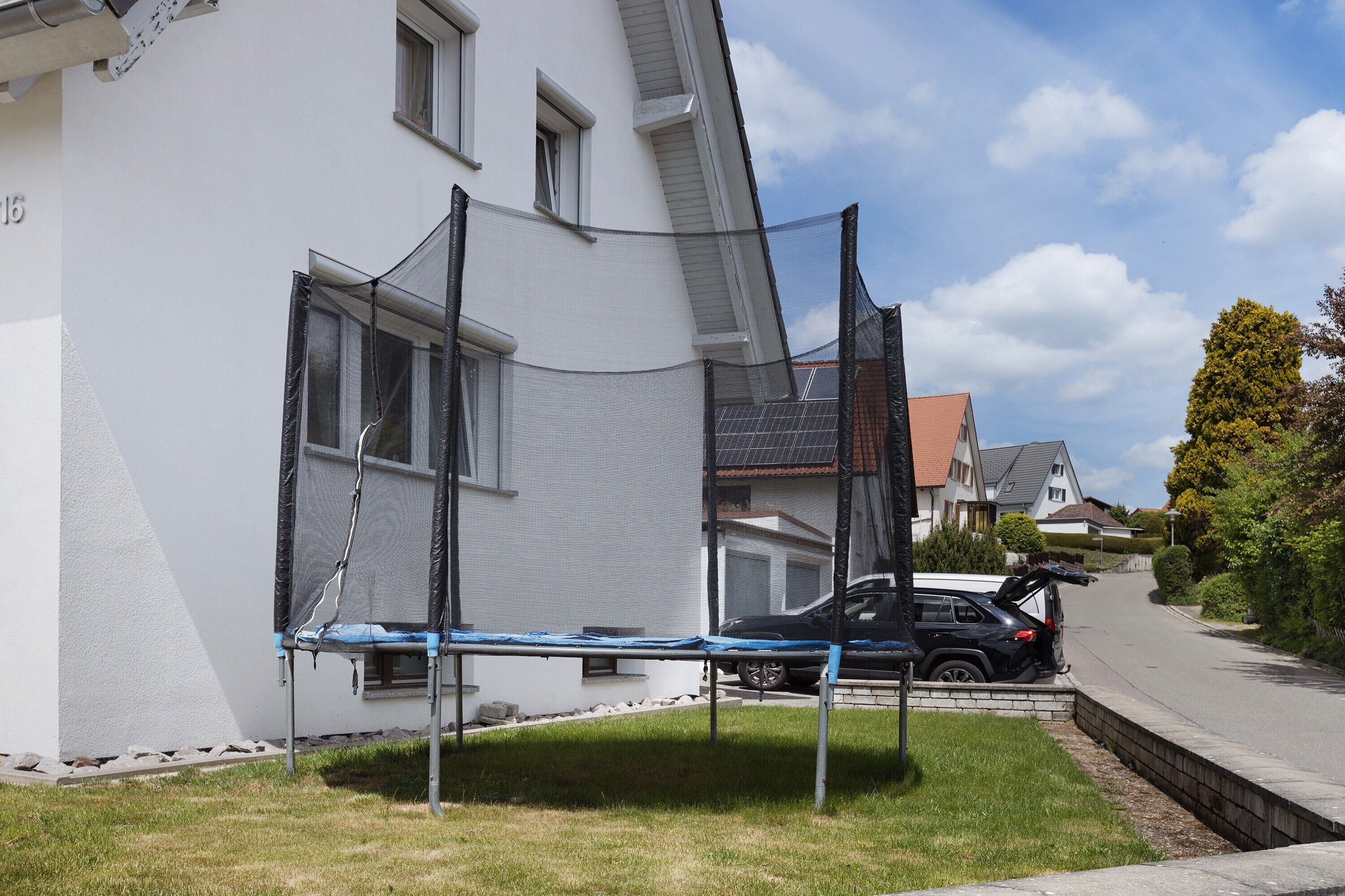 A residential street with a white house, a trampoline, and a parked car.