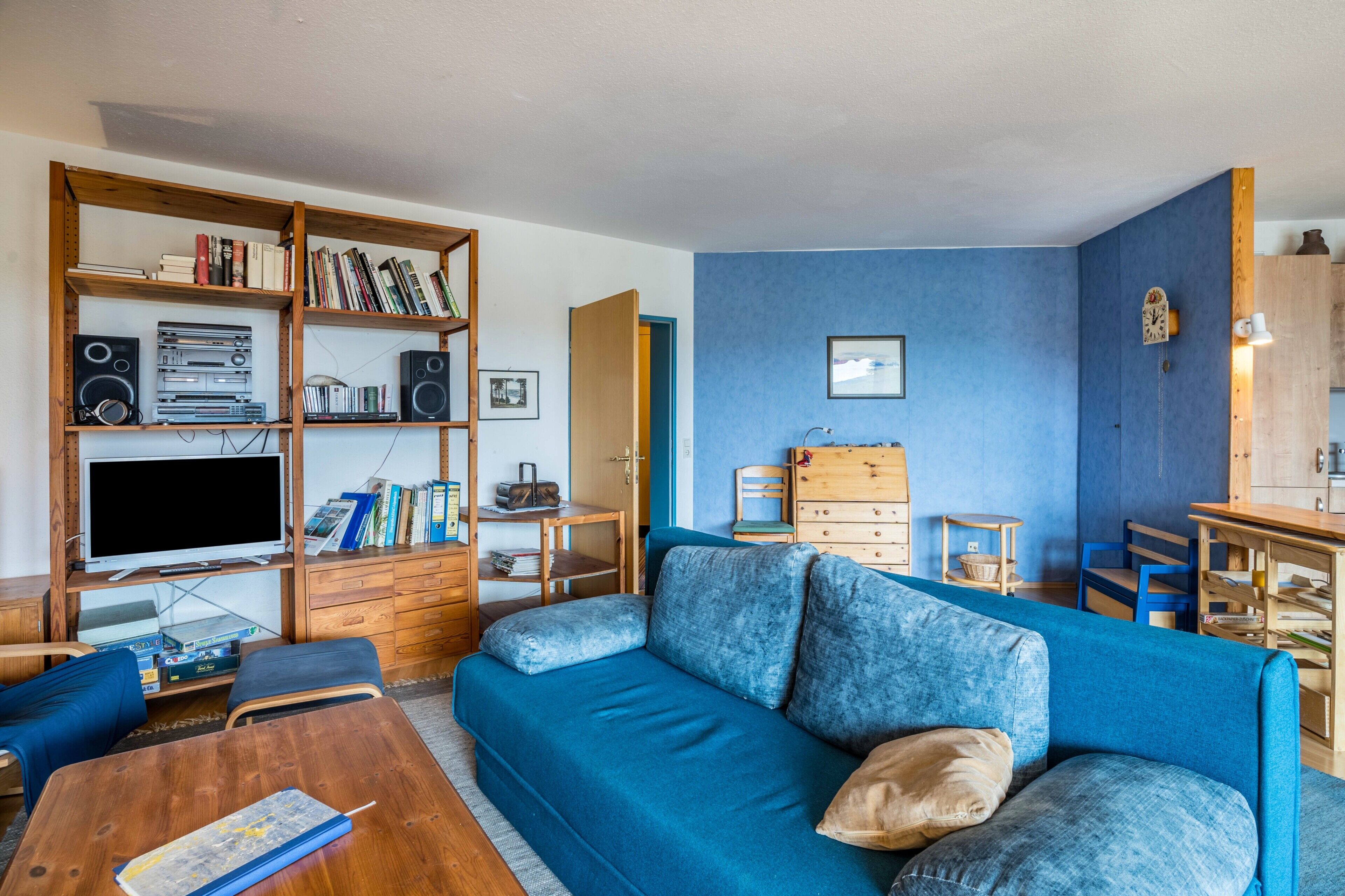 A living room with a blue sofa, wooden shelves with books and audio equipment, a television, and a wooden coffee table.