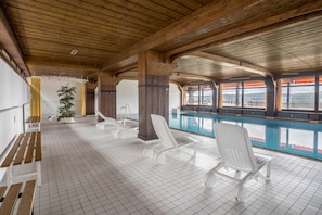 Area piscina con travi in legno, pavimento in piastrelle bianche e poltroncine da relax bianche.